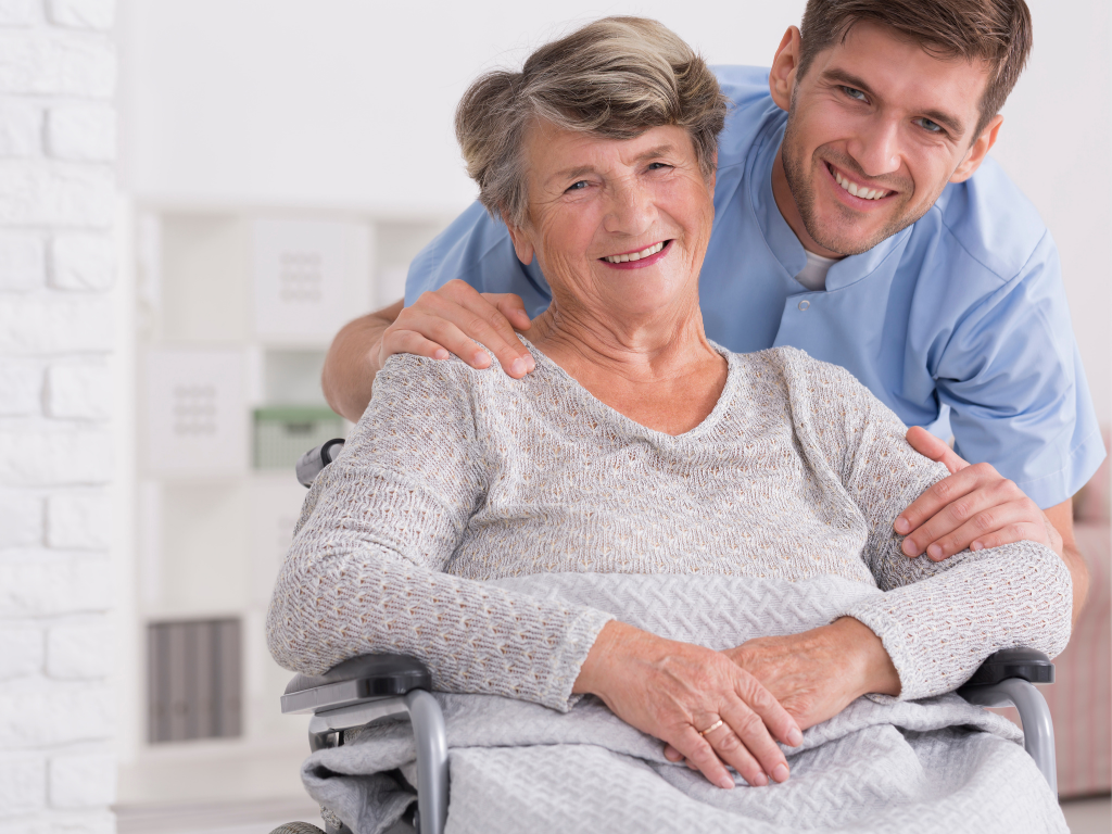 Smiling elderly woman in a wheelchair with a friendly male caregiver standing beside her, both looking at the camera in a bright home setting.. Senior home care in Barnstable County