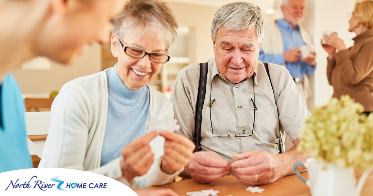 An older couple works on a puzzle with a caregiver, representing the kind of activity that helps those with dementia and also representing Alzheimer’s and Brain Awareness Month.