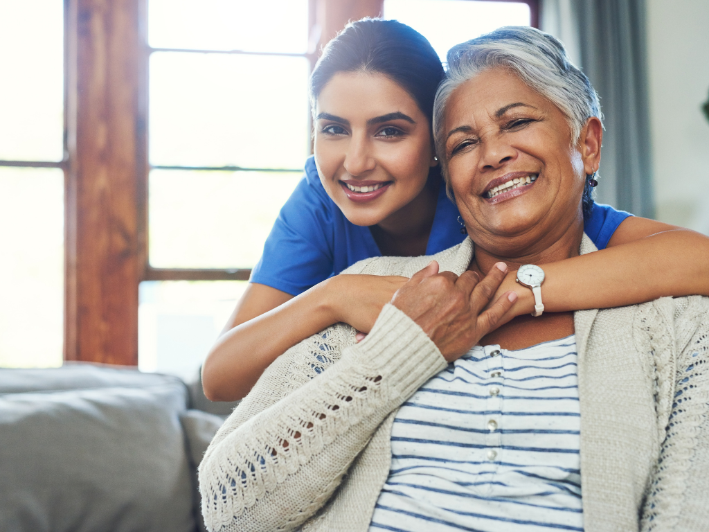 A smiling elderly woman sits comfortably on a couch while a cheerful caregiver in blue scrubs stands behind her, wrapping her arms around the senior in a warm, supportive embrace. The bright, cozy living room in the background emphasizes the caring environment. This image represents compassionate senior home care in Bourne, highlighting the strong bond and trust between caregivers and seniors.