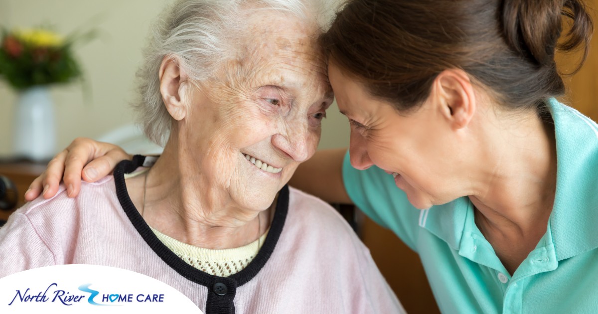 A caregiver lovingly embraces a senior woman while they both smile, representing the kind of warm disposition that can help when dealing with dementia-related repetition.