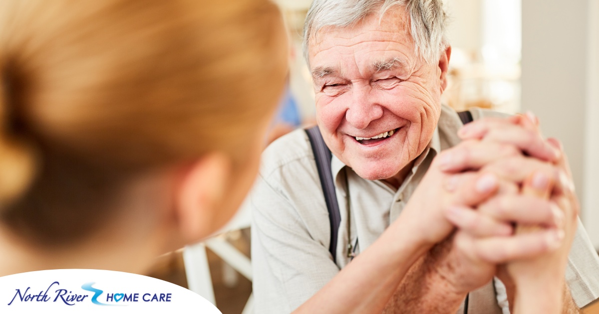 A caregiver holds hands with a happy older client, representing the results of creating a comfortable environment in the home.