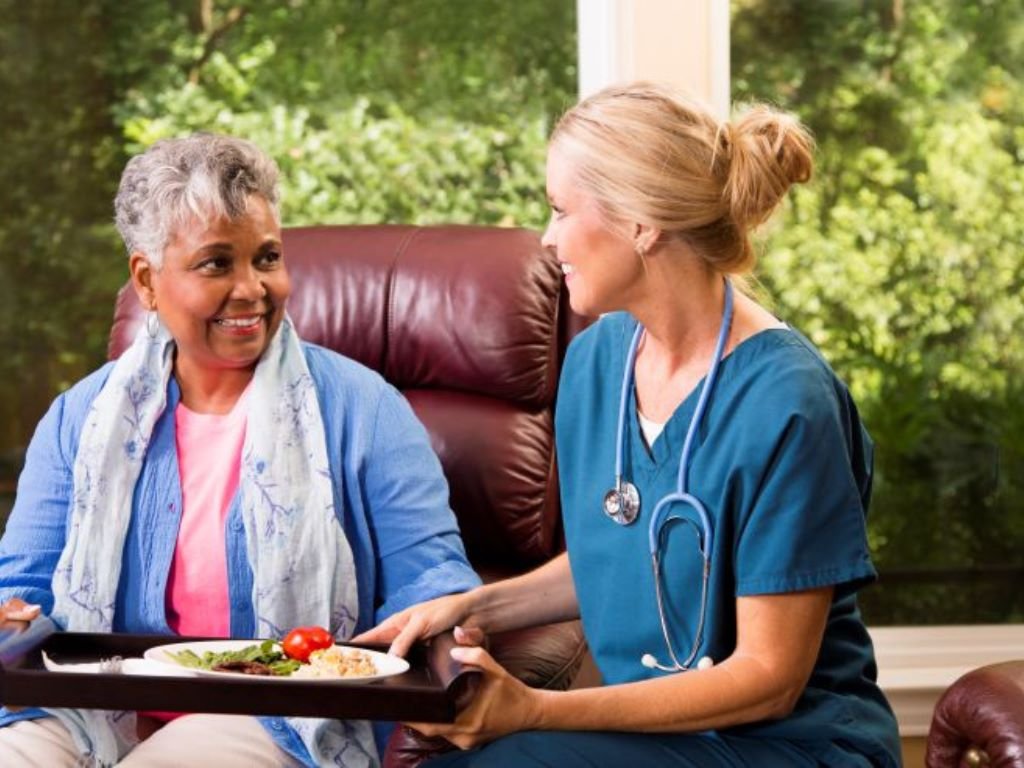 A care giver handing an elderly woman food sitting in a red chair. Home-Aide-Services-in-Abington-MA.png