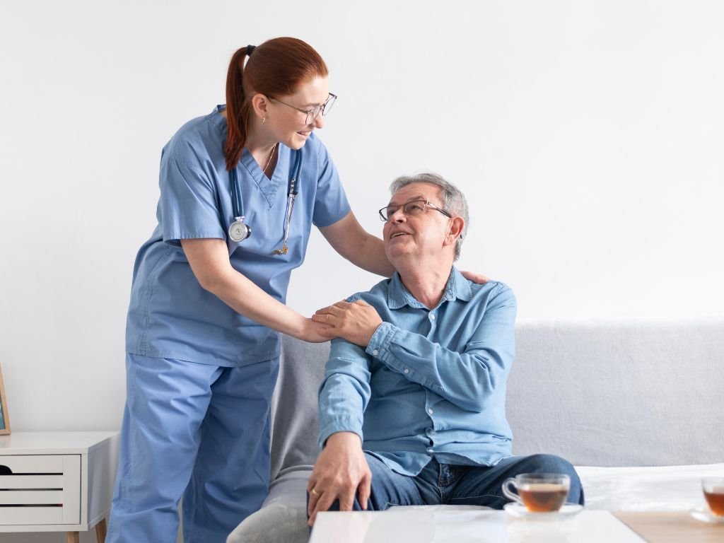 A young caregiver places her hands on the shoulder of a male senior who sits on a sofa, gently resting his hand on hers as they look at each other warmly. Home Care in Wellesley
