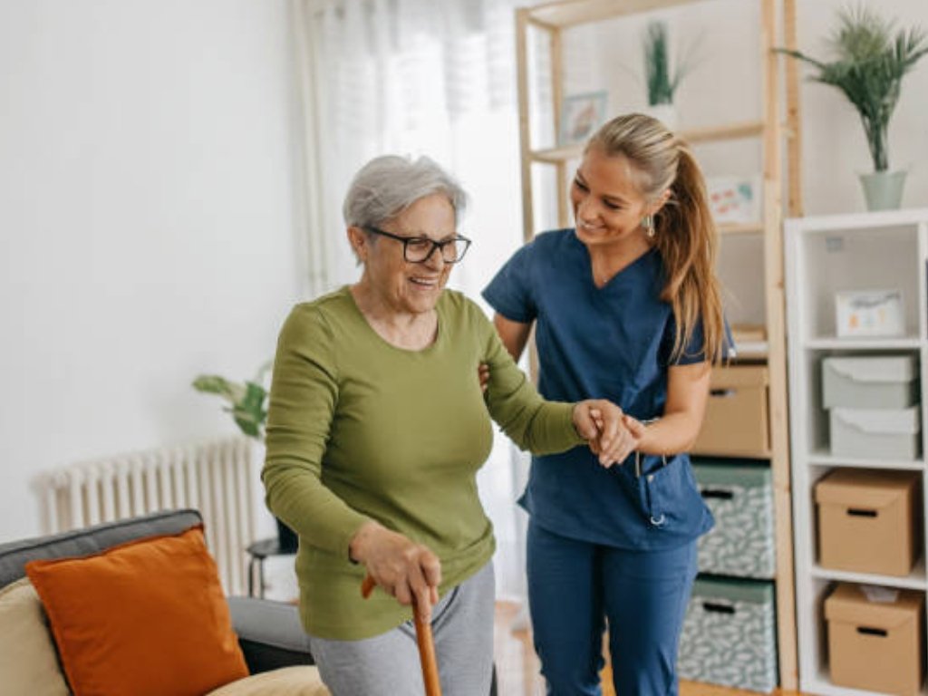 A smiling caregiver in blue scrubs assists an elderly woman with a cane inside a cozy, well-lit living room. The warm interaction reflects quality senior home care in New Bedford, emphasizing support, comfort, and independence for older adults.