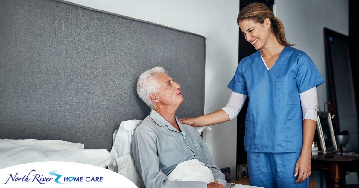 A caregiver smiles at an older client in bed, representing the compassion involved in being a live-in caregiver.